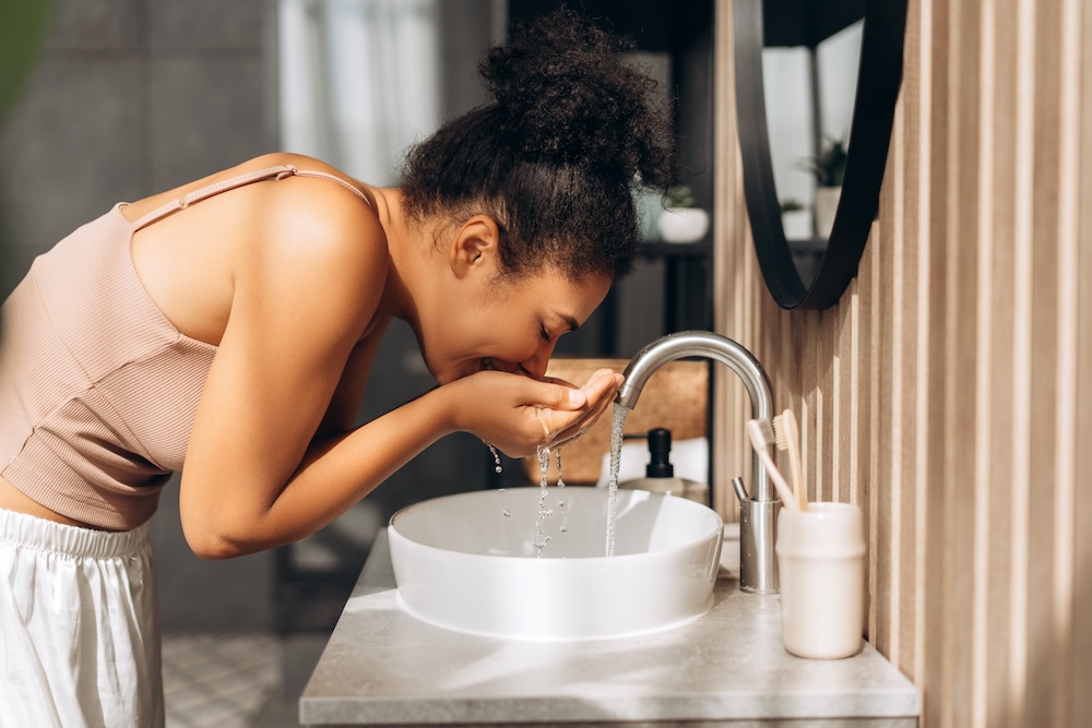 Frau beugt sich über ein Waschbecken und wäscht ihr Gesicht mit Wasser in einem Badezimmer.