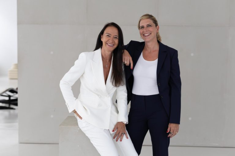 Zwei Frauen in eleganter Businesskleidung posieren selbstbewusst in einem hellen Studio; eine trägt einen weißen, die andere einen dunklen Hosenanzug.