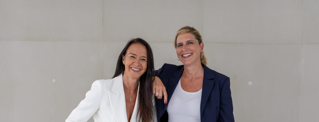 Zwei erwachsene Frauen stehen eng beieinander, lächeln herzlich und tragen helle Business-Outfits in einem modernen Innenraum.