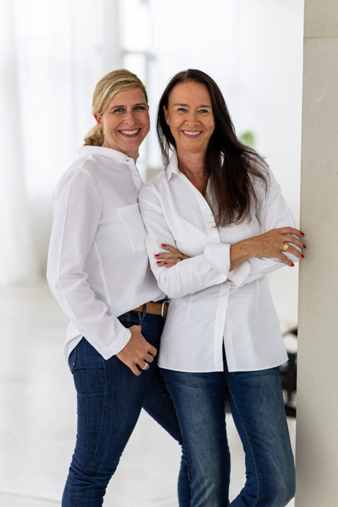 Zwei erwachsene Frauen stehen eng nebeneinander, lächeln in die Kamera und tragen helle, legere Business-Outfits vor neutralem Hintergrund.