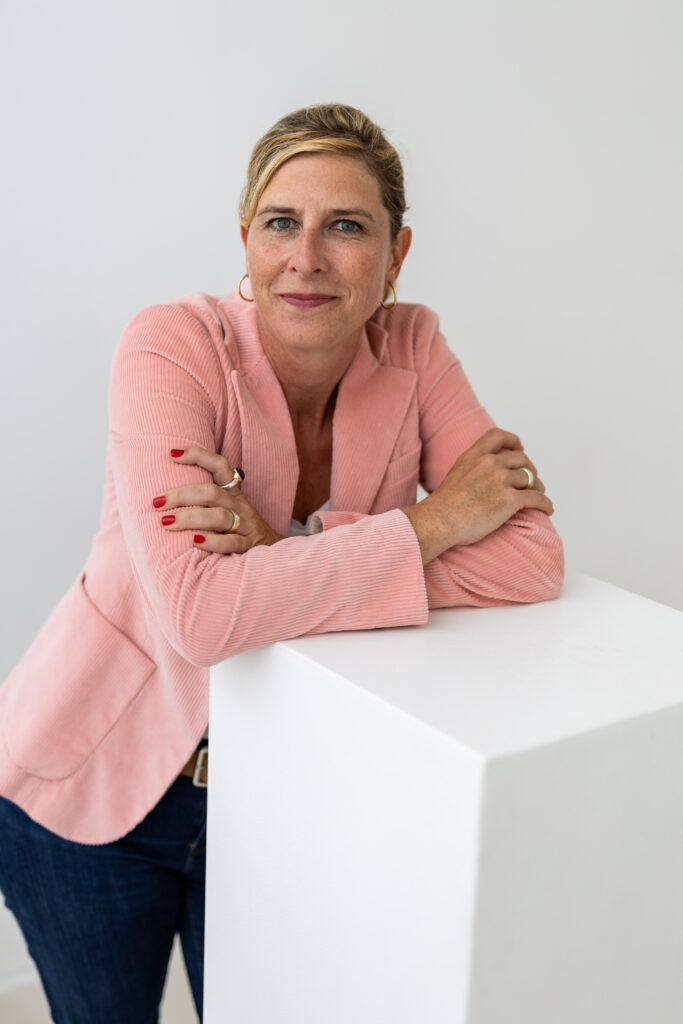 Frau im rosafarbenen Blazer lehnt an einem weißen Podest und blickt ruhig und direkt in die Kamera.