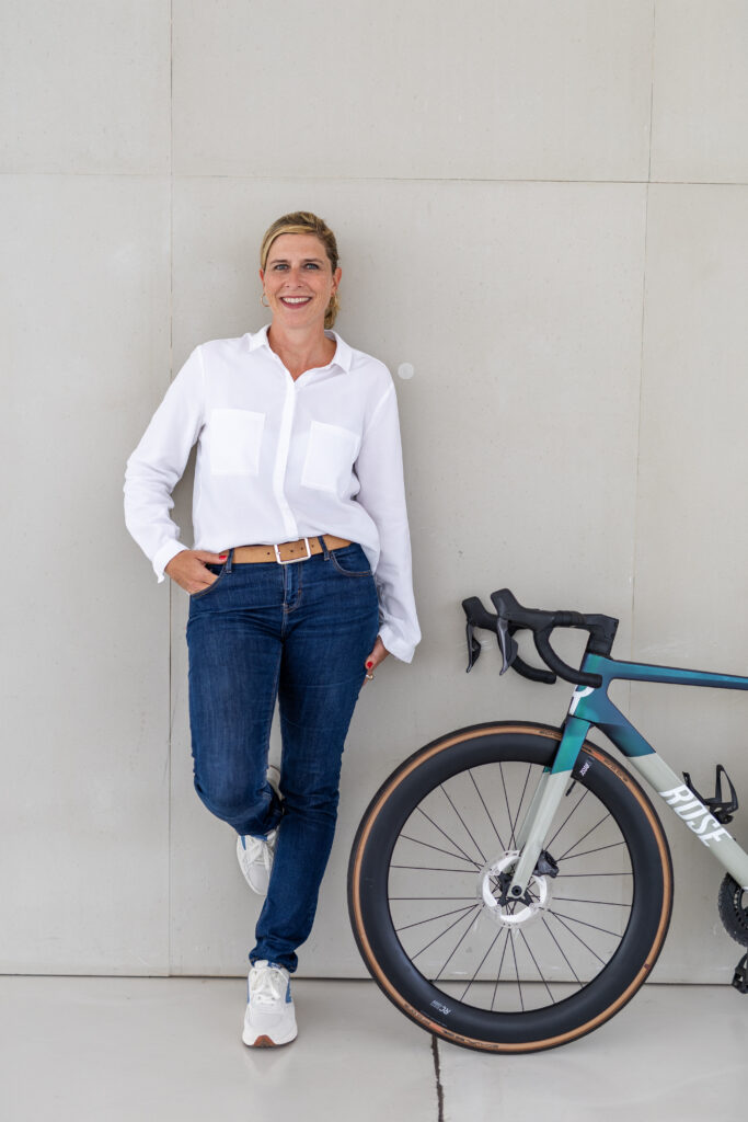 Frau in weißer Bluse und Jeans lehnt entspannt an einem Fahrrad in einem minimalistischen Innenraum.