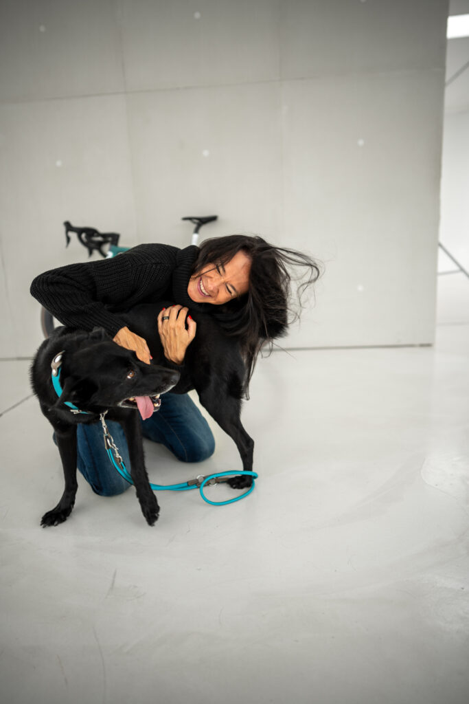 Frau sitzt entspannt auf dem Boden eines Studios und hält einen schwarzen Hund an der Leine; beide wirken ruhig und vertraut.