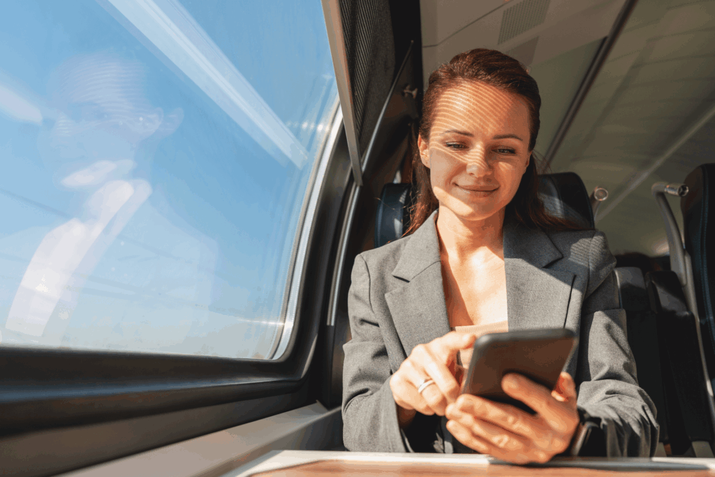 Geschäftsfrau sitzt am Fenster eines Zuges oder öffentlichen Verkehrsmittels und nutzt ein Smartphone bei Tageslicht.