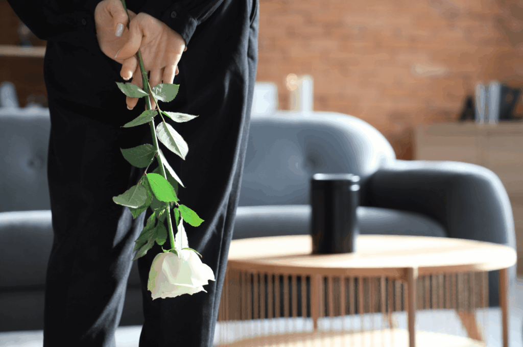 Hand einer Person, die eine weiße Rose kopfüber hinter dem Rücken hält; im Hintergrund ein modernes Wohnzimmer mit Sofa, Couchtisch und Becher, weich unscharf dargestellt.