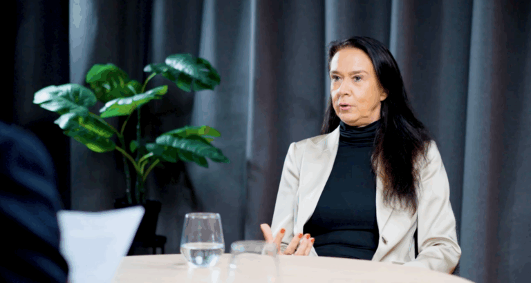 Petra Ahrens in einem Gesprächs- oder Interviewsetting, sitzend an einem Tisch, gestikulierend während des Sprechens; sie trägt ein helles Sakko über einem dunklen Oberteil, vor einem dunklen Vorhanghintergrund mit Zimmerpflanze und Wasserglas im Vordergrund.