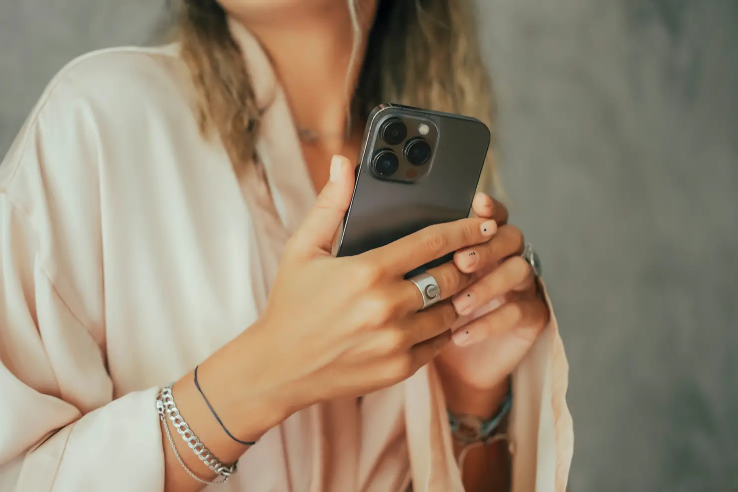 Person hält ein modernes Smartphone in beiden Händen; Nahaufnahme der Hände mit Schmuck und hellem Outfit.