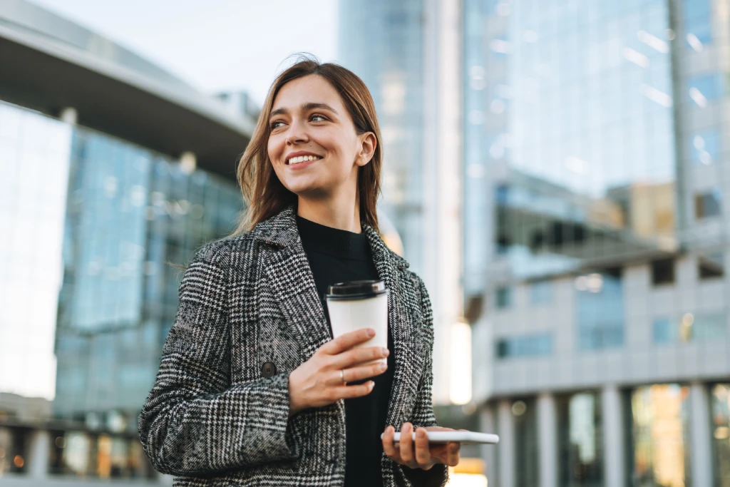 Geschäftsfrau steht im urbanen Umfeld mit einem Coffee-to-go-Becher und lächelt entspannt.