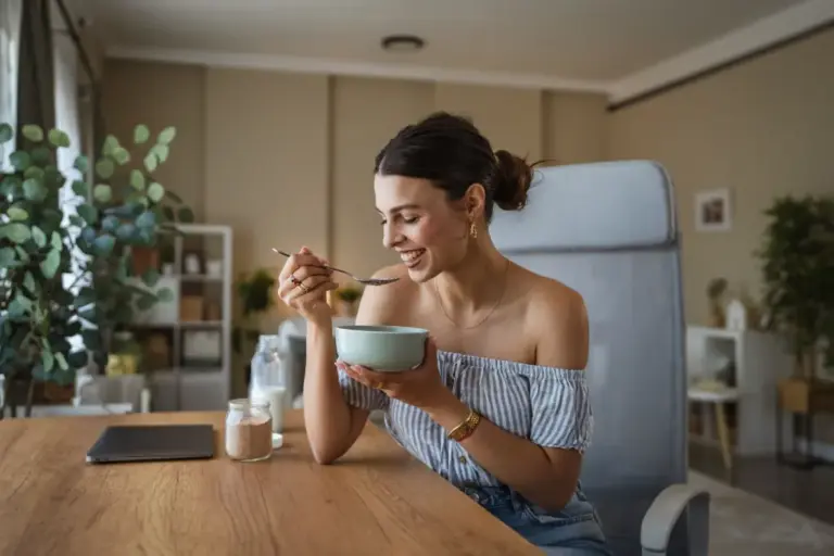 Junge Frau sitzt entspannt auf einem Sofa und isst lächelnd eine Schüssel Joghurt oder Frühstücksbrei in einem hellen Wohnraum.