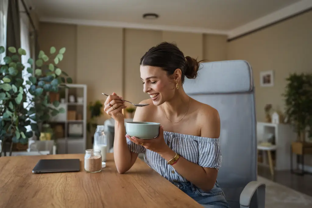 Junge Frau sitzt entspannt auf einem Sofa und isst lächelnd eine Schüssel Joghurt oder Frühstücksbrei in einem hellen Wohnraum.