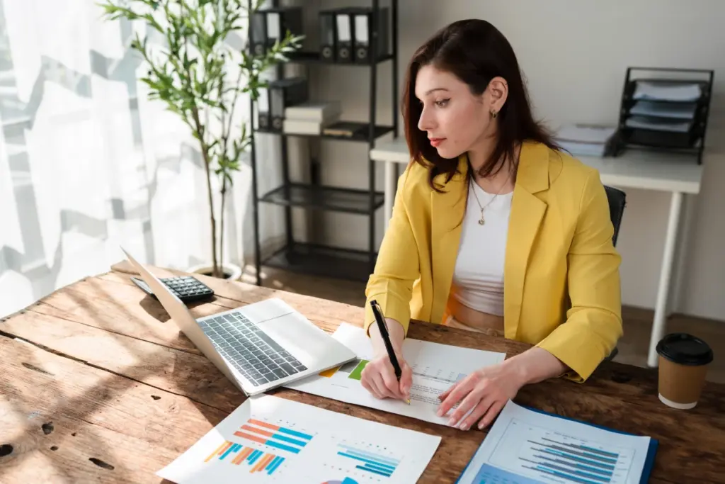 Frau analysiert Finanzunterlagen und Diagramme am Schreibtisch mit Laptop und Notizen – Financipation Club