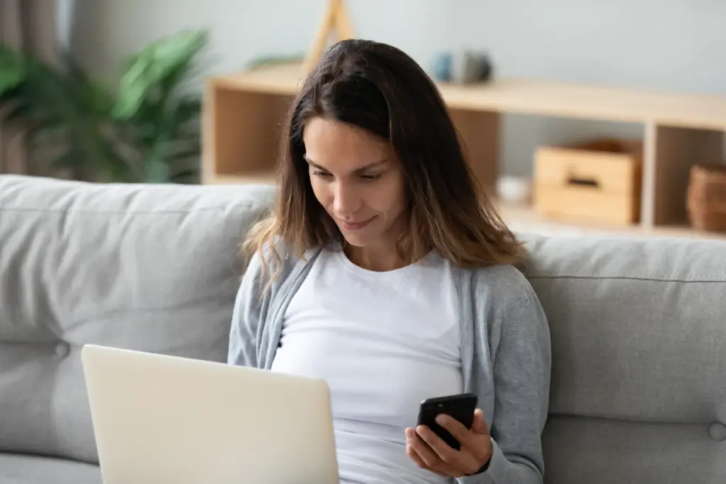 Junge Frau sitzt zu Hause auf dem Sofa und nutzt gleichzeitig Laptop und Smartphone – Financipation Club